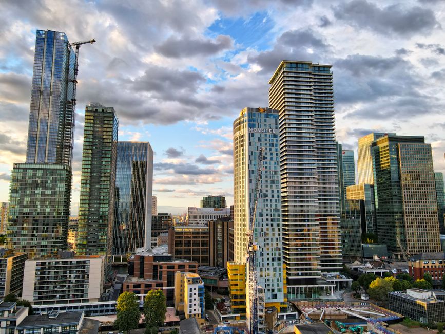 A wide shot of the Canary Wharf area during daytime showing tall modern office buildings and residential apartments lining the riverfront. Visible are high-rise structures with glass facades, includin